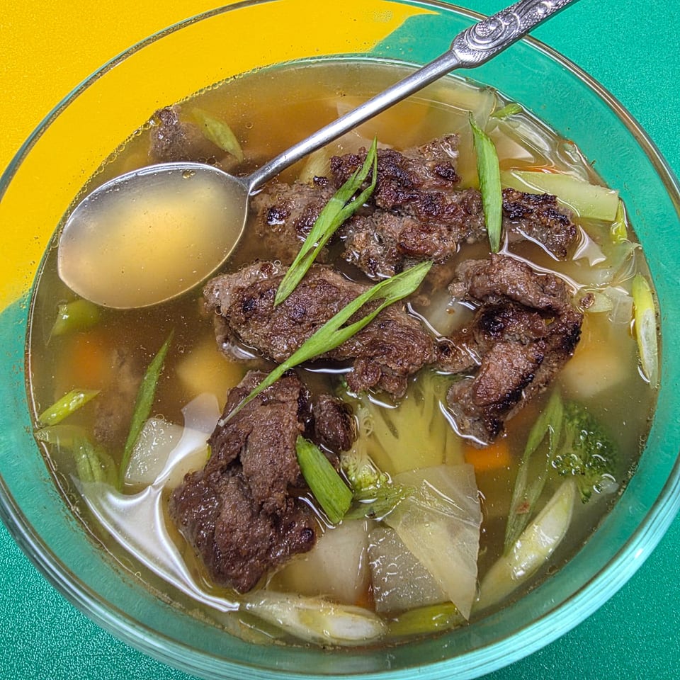 A clear bowl of beef and vegetable soup with a spoon, on a yellow and green background.