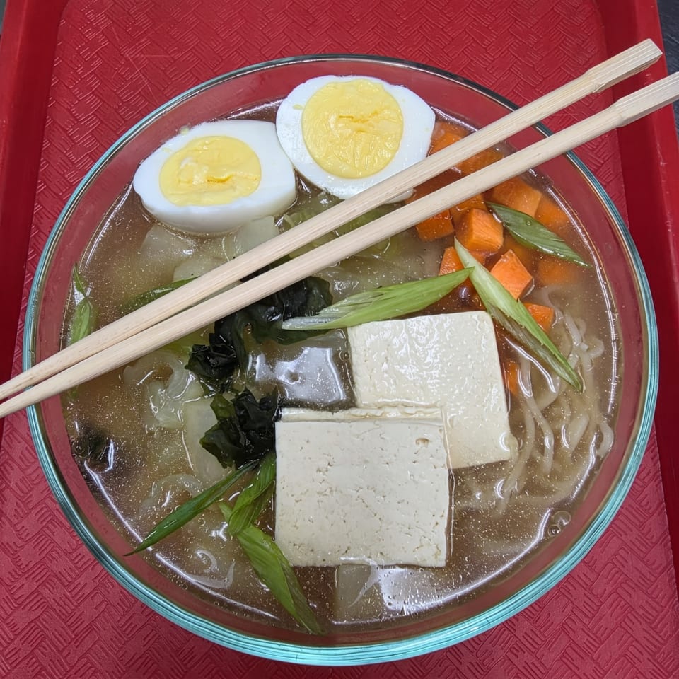 Ramen bowl with noodles, broth, tofu, hard-boiled eggs, carrots, and green onions, with chopsticks.